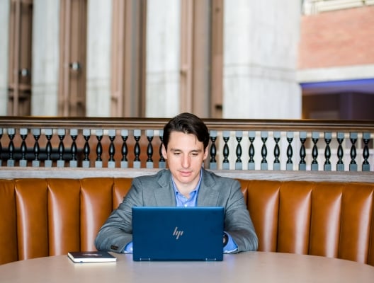 A man in a suite sitting in a leather bench working on a laptop