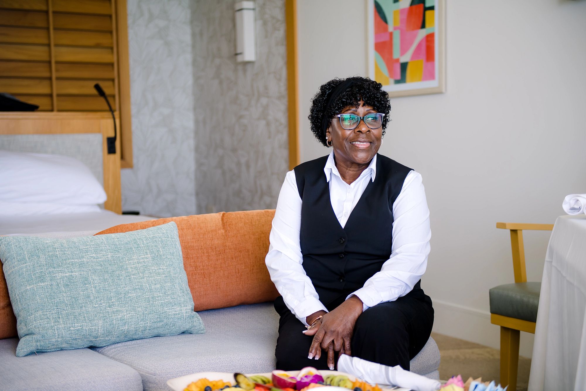 A gentleman of a visual minority dressed in business clothing. She is sitting on the edge of a couch.