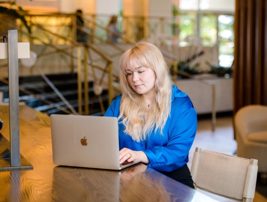 A blonde woman in a professional outfit, sitting at a hotel desk working on a laptop