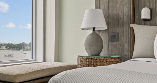 Room at Newport Harbor Island Resort with a bench in front of a window over looking the water. In the foreground is a bed with a bedside table with a lamp.